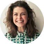 A woman with curly brown hair smiles broadly, wearing a green and white patterned shirt, standing against a plain light-colored background.