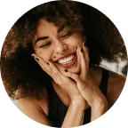 A woman with curly hair smiles with her eyes closed, resting her face in her hands. She wears a sleeveless top and has black nail polish.