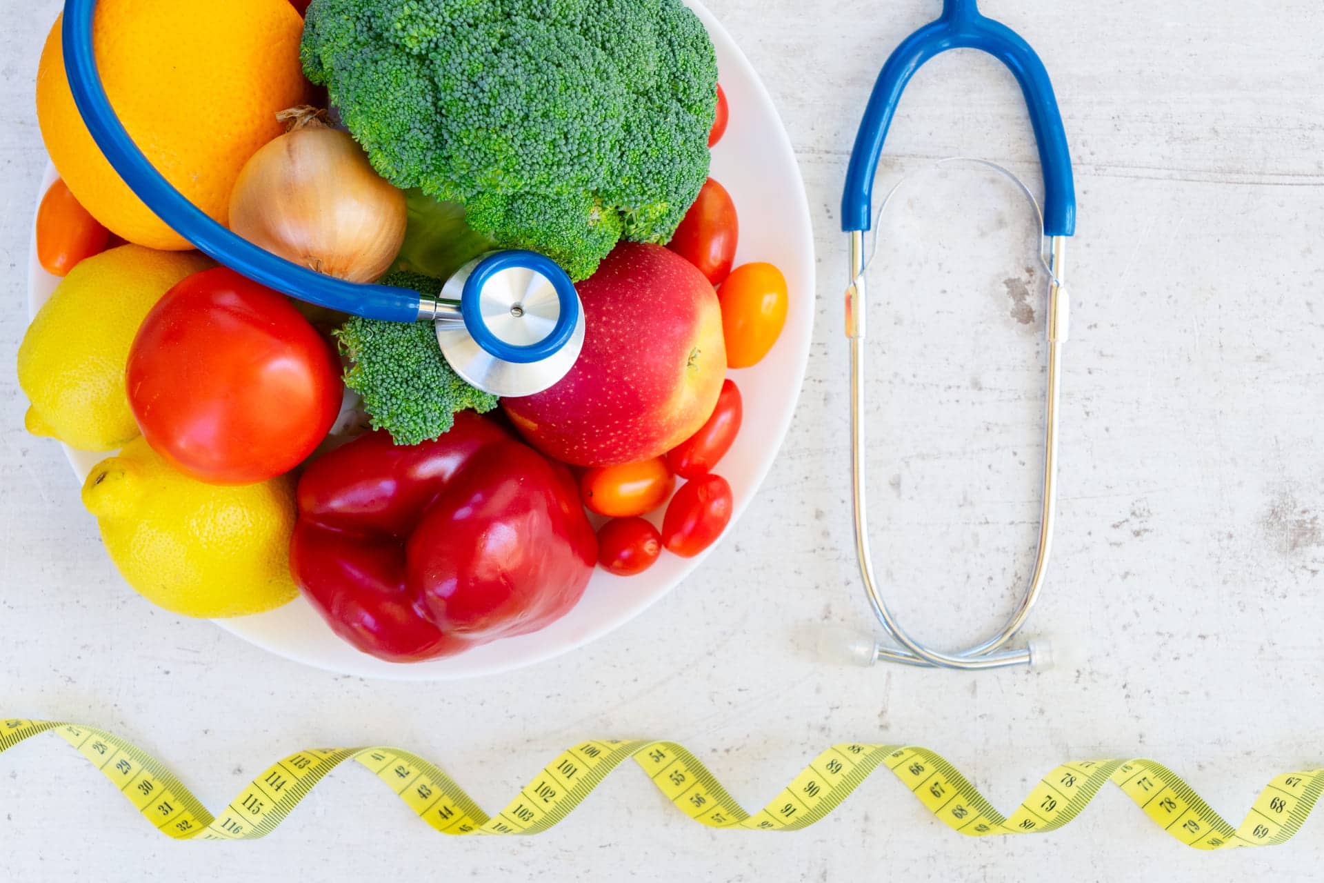 A bowl of fresh fruits and vegetables with a stethoscope, a yellow measuring tape, and a white background.