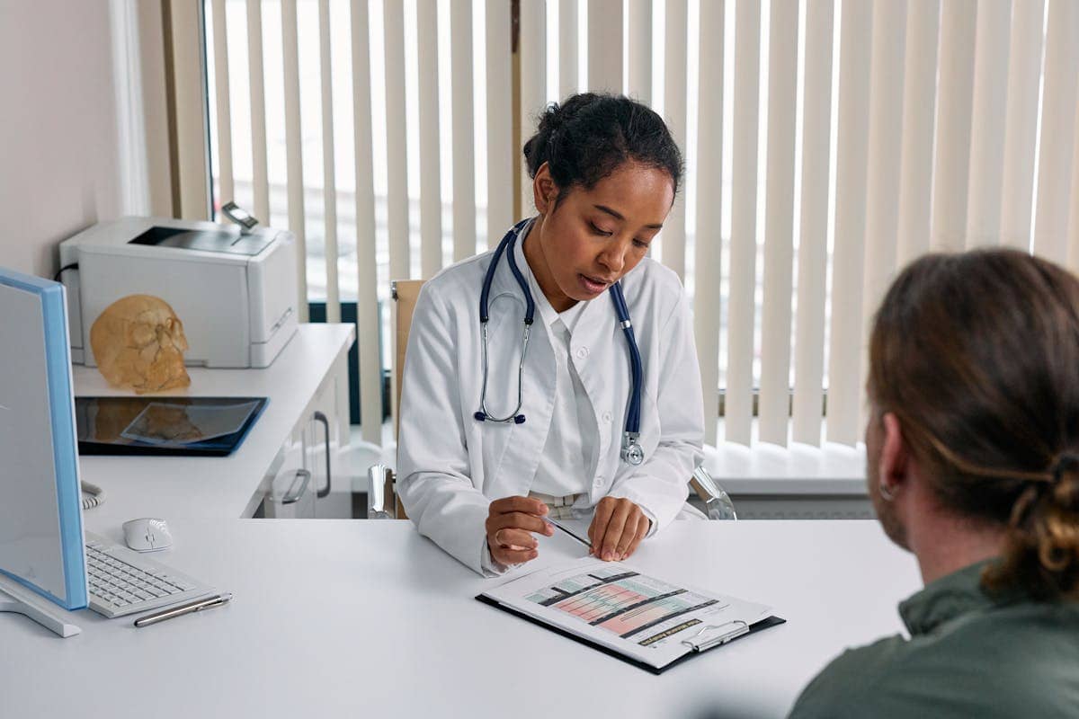 A doctor in a white coat reviews a medical chart with a patient seated across a desk in a well-lit office.